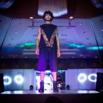 Modelo masculino desfila com um look de moda em uma passarela iluminada.