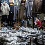 Interior movimentado de uma loja de roupas, com funcionários organizando produtos e cuidando do estoque.
