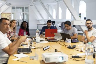 Equipe diversa reúne-se em torno de uma mesa de conferência, usando laptops e discutindo projetos em um escritório moderno.
